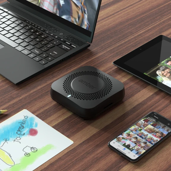 An Amber X device on a tabletop surrounded by a laptop, phone, and tablet.